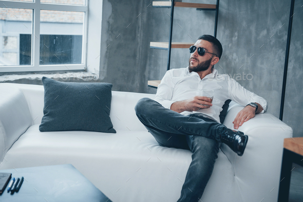 Thoughtful serious look. Young short-haired man in sunglasses sitting on couch in the office
