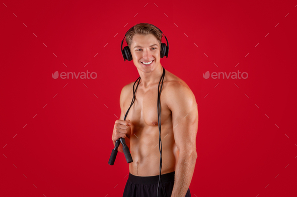 Attractive shirtless young guy with skipping rope listening to music in headphones on red studio