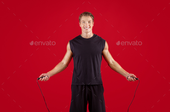 Cardio workout concept. Young fit sportsman jumping on skipping rope over red studio background