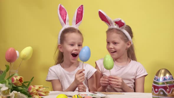 Happy Little Girls with Bunny Ears Smiling and Demonstrating Painted Eggs on Sticks While Sitting at