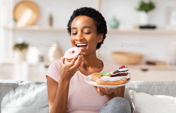 Young black woman on diet having cheat meal day, holding plate of sweets, stuffing her mouth with