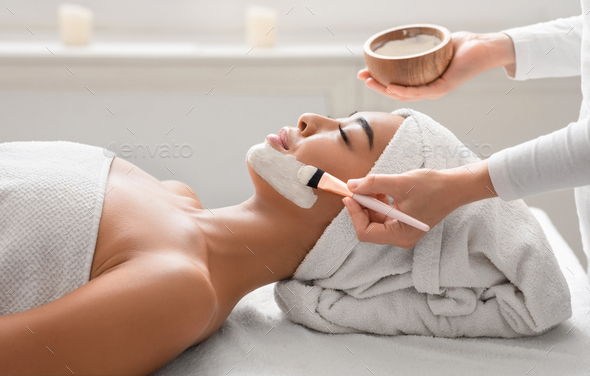 Relaxed asian woman getting facial nourishing mask by beautician at cosmetology cabinet