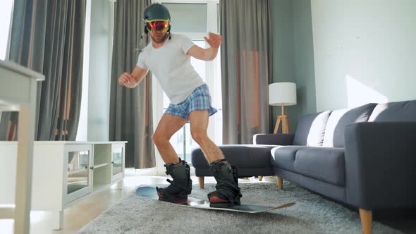 Fun Video. Man in Shorts and a T-shirt Depicts Snowboarding on a Carpet in a Cozy Room. Waiting for alt