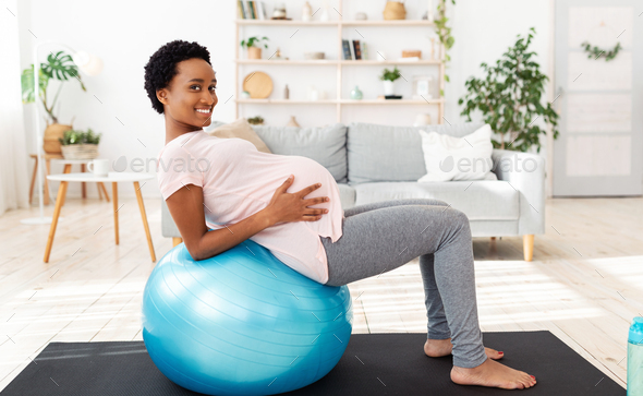 Exercising during pregnancy concept. Positive black future mother working out on fitness ball at
