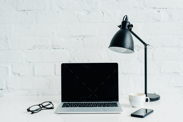 Laptop with blank screen, smartphone, lamp and eyeglasses on desk