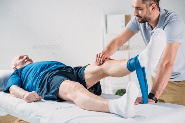 rehabilitation therapist assisting senior man exercising with rubber tape on massage table