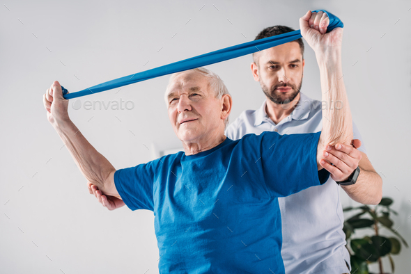 focused rehabilitation therapist assisting senior man exercising with rubber tape