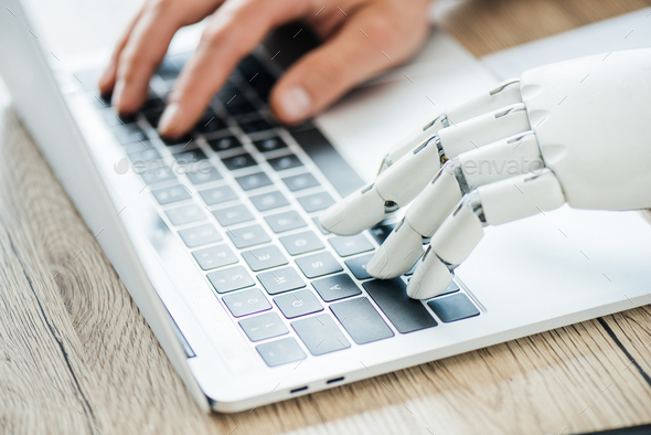 close-up view of human and robot hands typing on laptop at workplace