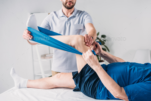 cropped shot of rehabilitation therapist assisting senior man exercising with rubber tape on massage