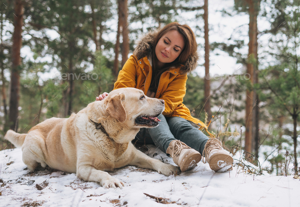 Young smiling woman in yellow jacket with big kind white dog Labrador walking in winter forest