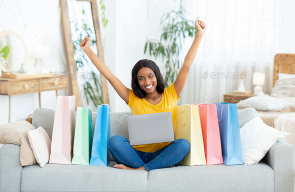 Excited black lady shopping online, sitting on sofa with laptop computer, surrounded by gift bags