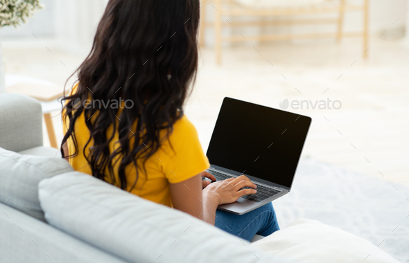 Unrecognizable black lady typing on laptop with blank screen on comfy couch at home, mockup for