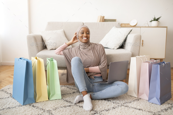 Happy black lady in hijab sitting on floor with laptop and paper bags, shopping on internet at home