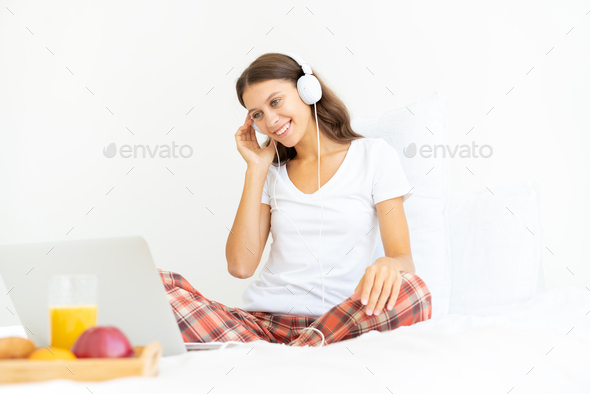 Young smiling woman sitting on bed in bedroom and listening to music or watching movie