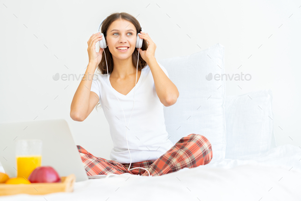 Young smiling woman sitting on bed in bedroom and listening to music