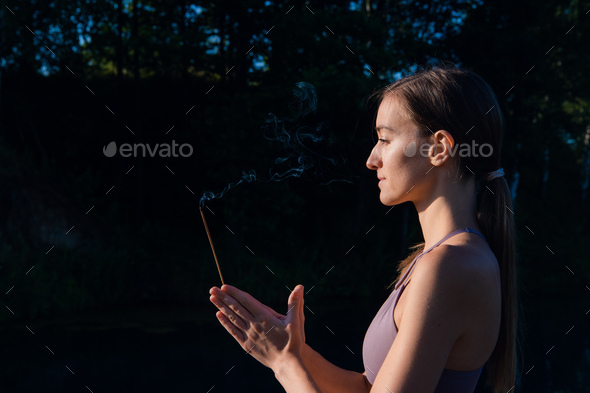 Yoga woman in sport clothes meditate and breathe on a sunrise in nature. Smoke from aromatic stick in hands
