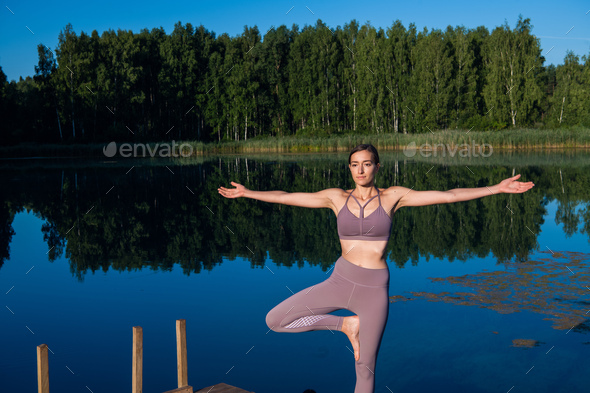 Young woman by the lake practicing yoga moves on wooden platform. Pretty young woman exercising in nature, healthy lifestyle young people positive vibes