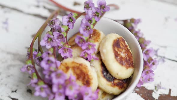 A Bouquet of Lilac Flowers Near a Plate with Cheese Cakes on a Wooden Background alt
