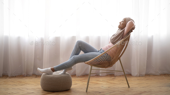Black Muslim woman in hijab relaxing in lounge chair at home, putting her feet on pouf, closing eyes