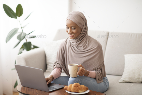 Online work, education, communication concept. Happy black lady in hijab using laptop and drinking