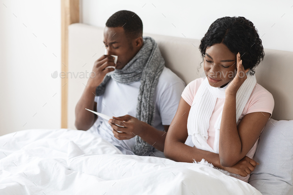 Sick african couple sitting in bed, measuring fever, sneezing noses