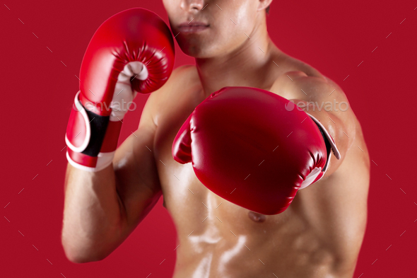 Determined young boxer in fighting gloves making punch at camera on red studio background, selective