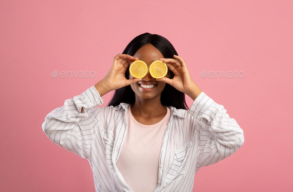Happy young black lady with lemon halves in front of her eyes posing on pink studio background