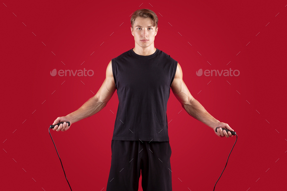 Aerobic workout concept. Serious athletic millennial guy jumping on skipping rope over red studio