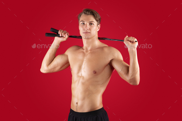 Motivated young guy with sexy body holding skipping rope on red studio background