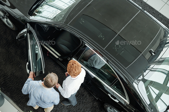 View of young salesman of auto center showing car interior to mature male client