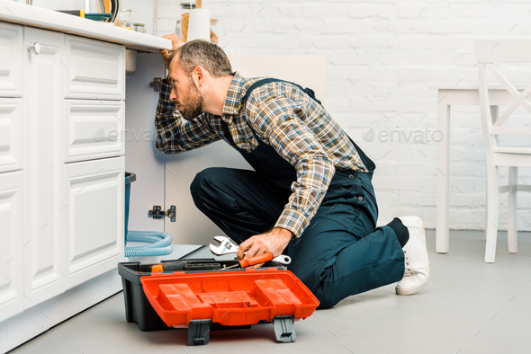 handsome plumber holding adjustable wrench and looking under broken sink in kitchen