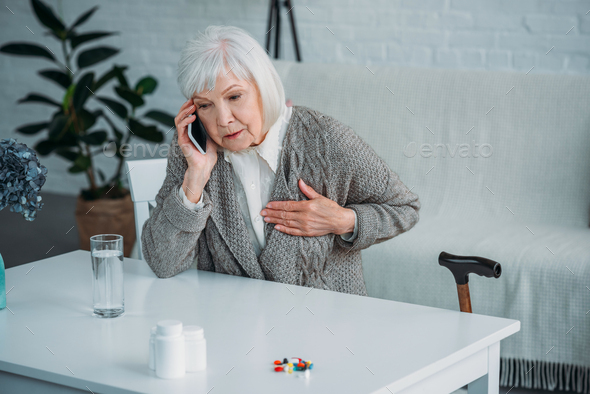 portrait of senior woman with heart ache talking on smartphone at table with medicines at home