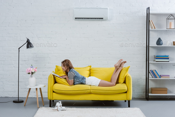 beautiful young woman reading book on sofa under air conditioner hanging on wall