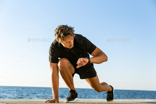 handsome adult sprint runner looking at fitness tracker while standing in start position for run on
