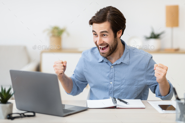 Excited Young Businessman At Laptop Computer Shaking Fists In Office
