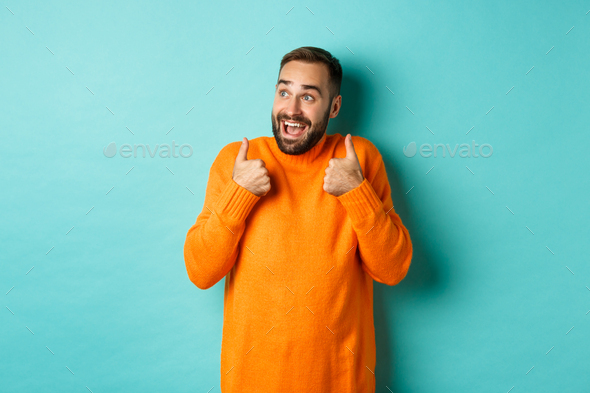 Cheerful man praising and looking at upper left corner, showing thumbs up amazed, super cool product