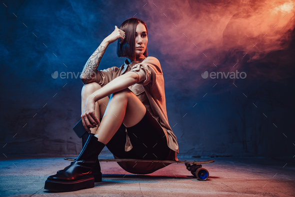 Hipster girl with tattoo and phone sits on skate in smokey