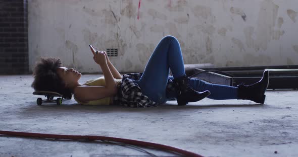 Mixed race woman in an empty building lying on a skateboard using smartphone laughing alt