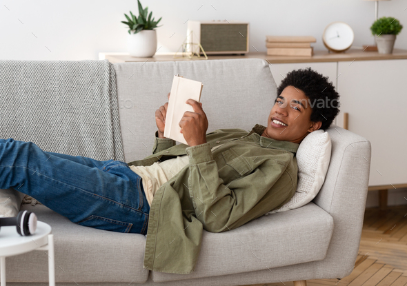Remote studies concept. Smart black teenager reading textbook on comfy couch at home