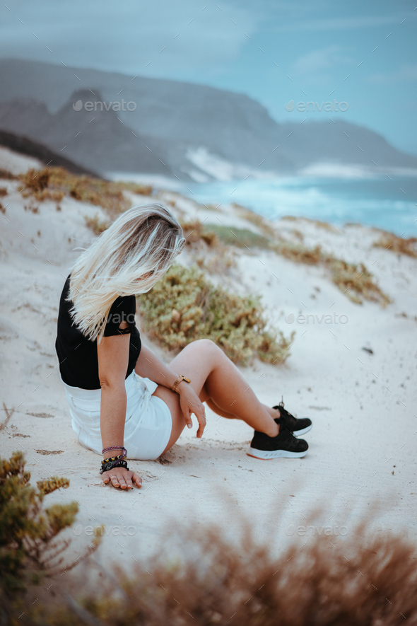 Young woman sitting on white sand dune surrounded by barren desert vegetation. Atlantic ocean - Stock Photo - Images