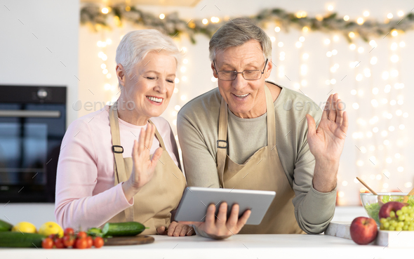 Senior Couple Waving Hello To Tablet Making Video Call Indoors