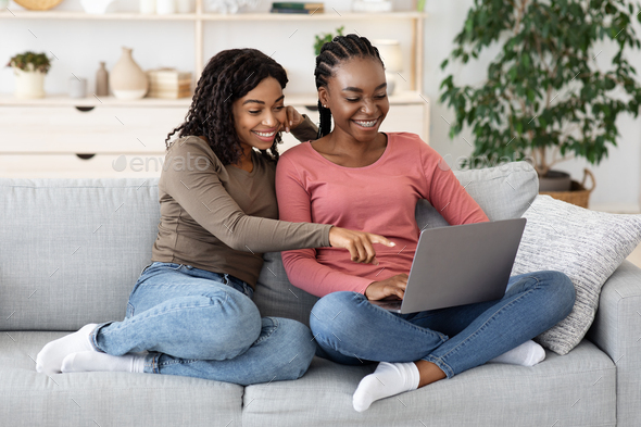 Two smiling black women using laptop at home, websurfing