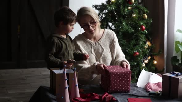 Mother with son wraps a Christmas gifts on a table alt