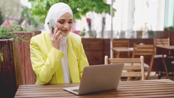 Smiling Young Muslim Woman in Headscarf Talking on the Phone and Using Laptop in Cafe alt
