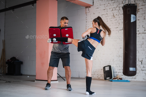 A young strong kickboxer girl kicks her sparring partner with a boxing paw in a personal training