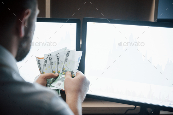 Bearded man in white shirt holds earned money in the office with multiple computer screens