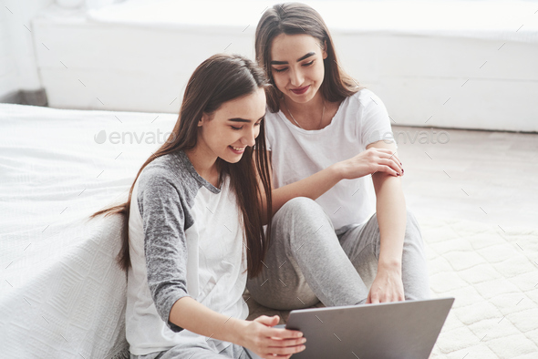 Young female twins sitting on the floor near white bed and using silver colored laptop