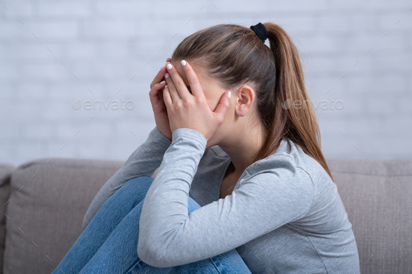 Side view of young woman with depression covering her face and crying on sofa at home