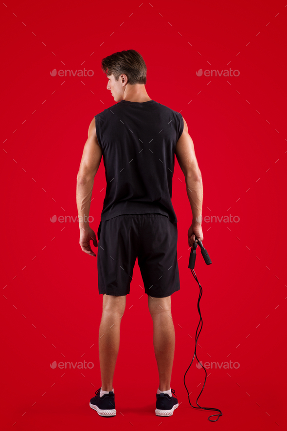 Aerobic workout. Back view of strong young man standing with skipping rope on red studio background