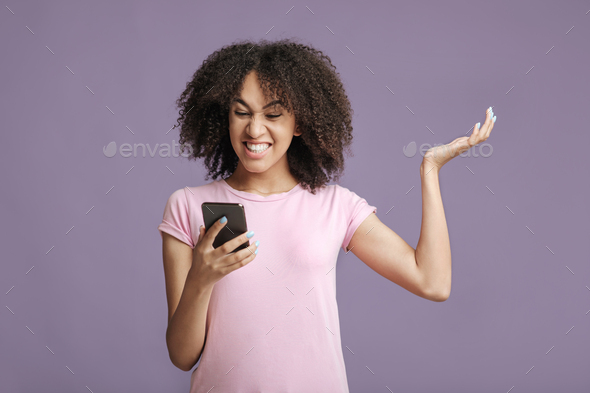 Upset millennial african american lady in pink t-shirt with curly hair, reads message and gets angry